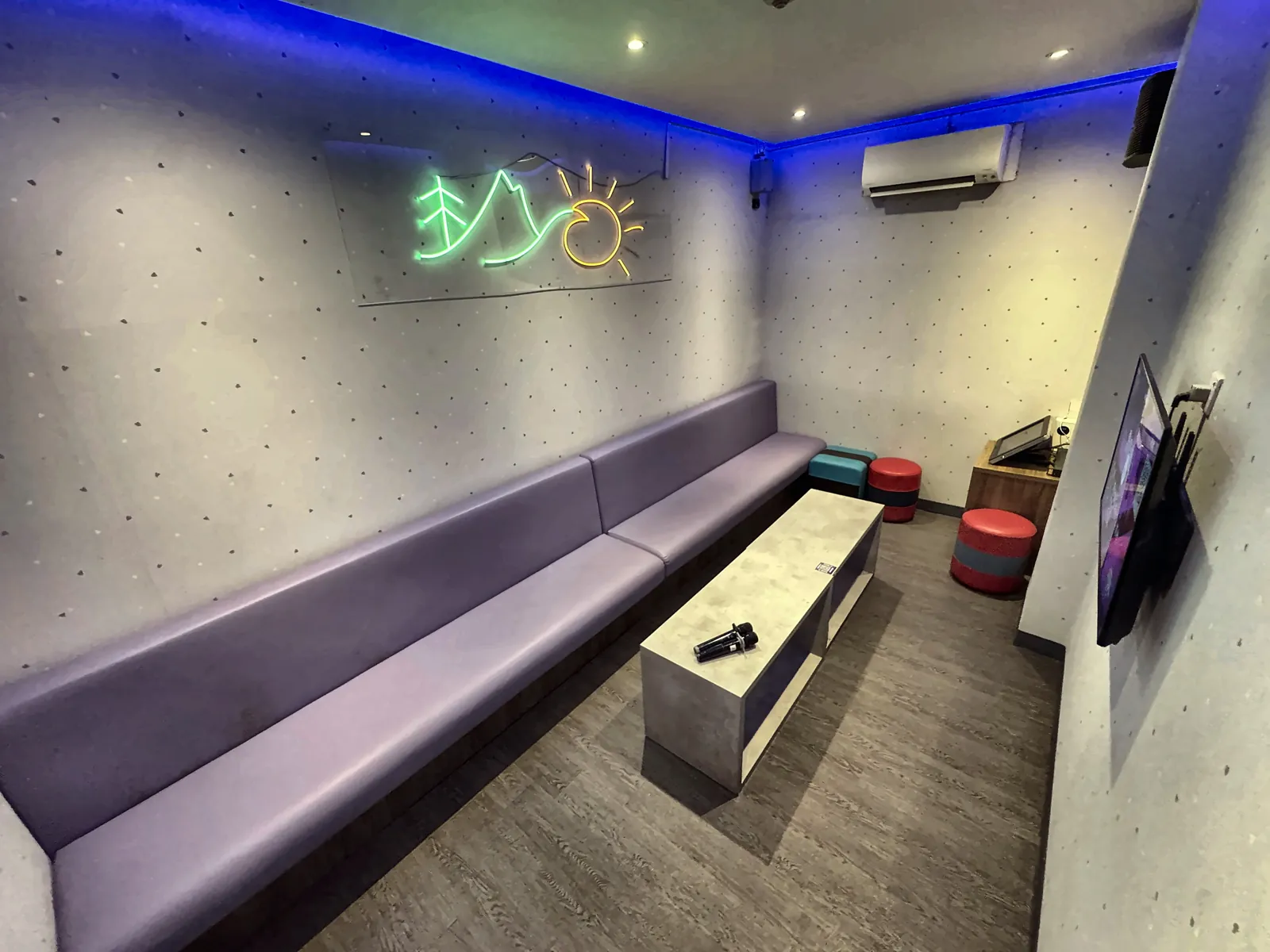 A private karaoke room at Major 99 with a long purple sofa, a coffee table with microphones, a wall TV and a neon sunrise-and-pine sign